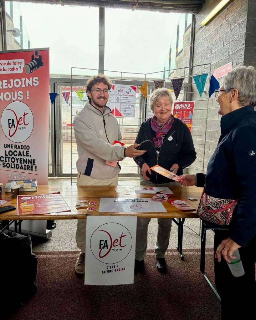 Un stand d'information pour la radio locale FA Jet, avec un homme en train de distribuer des brochures à deux femmes, dans un espace intérieur.
