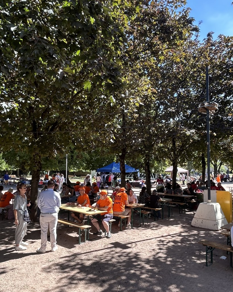 Un parc animé avec des tables sous des arbres, des gens assis en groupes, certains portant des t-shirts orange, une tente bleue au fond.