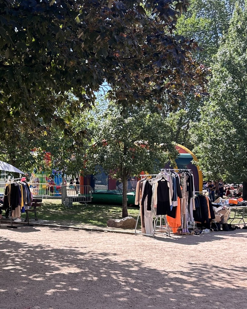 Un marché en plein air avec des vêtements exposés sur des portants, entouré d'arbres et d'une structure gonflable colorée en arrière-plan.