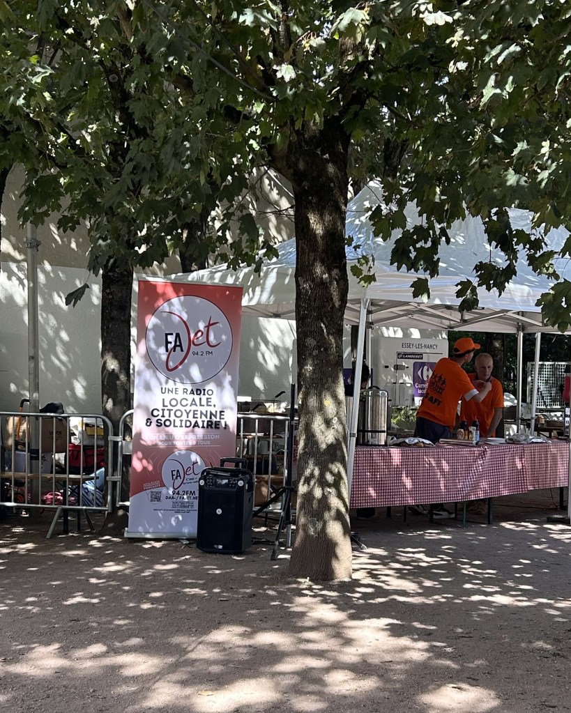 Un stand extérieur avec une pancarte pour la radio FAjet 94.2 FM, entouré d'arbres. Deux personnes en t-shirts orange interagissent près d'une table avec des boissons.