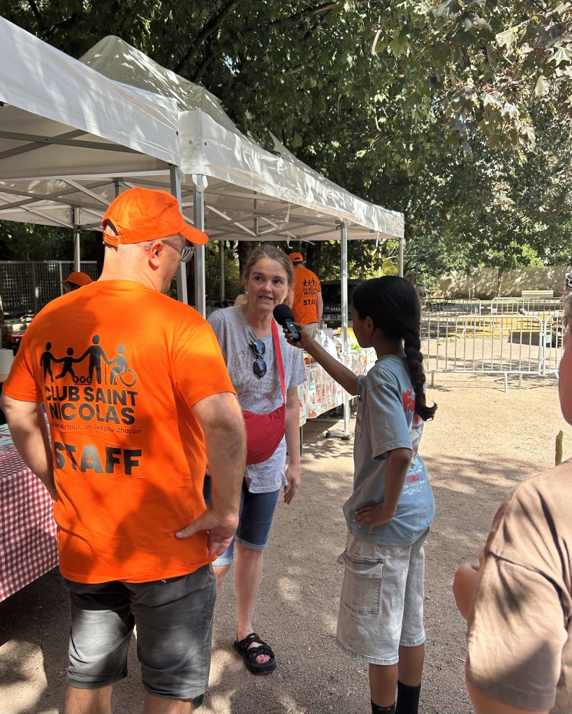 Un membre du personnel en t-shirt orange du Club Saint Nicolas discute avec une femme pendant qu'un enfant l'interroge, le tout sous des tentes au parc.